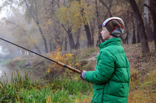 A child of 9 years old in a green jacket and with a fishing rod in his hands in the morning autumn fishing, river fog. Close-up. Blurred background.