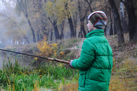 A child of 9 years old in a green jacket and with a fishing rod in his hands in the morning autumn fishing, river fog. Close-up. Blurred background.