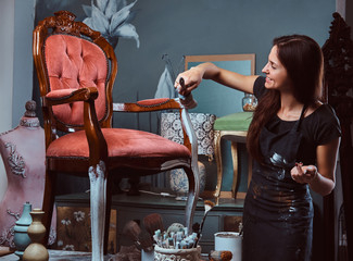 Female artist painting vintage chair in white color with paintbrush in workshop.