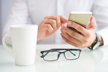 Close-up of man using smartphone at table with mug and glasses. Unrecognizable businessman viewing...
