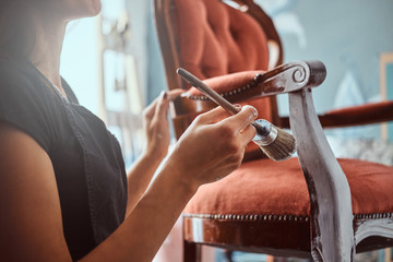 Female artist painting vintage chair in white color with paintbrush in workshop.