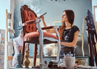 Female artist painting vintage chair in white color with paintbrush in workshop.