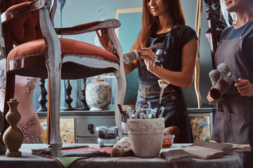 Female artist with her assistant in aprons painting vintage chair with paintbrush in workshop.