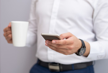 Close-up of young businessman using mobile phone indoors. Caucasian man standing with tea cup and reading or texting message on smartphone. Mobile communication concept