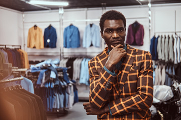 Elegantly dressed African man posing with hand on chin while standing in a classic menswear store.