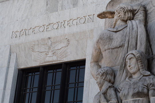 Text and an eagle carved into marble above the entrance to the Oregon State Capitol building in Salem, part of a marble relief carving representing white settlers in the foreground.