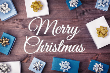 On a dark wooden background the inscription Merry Christmas. Blue and white gift boxes with decorative bows are laid out around it.