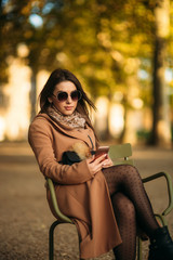 A young girl in a brown coat sits in the middle of the park on a khaki stool. Attractive girl use phone