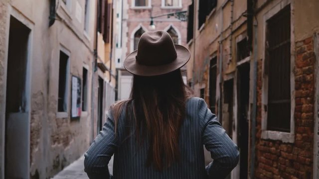Back view of confident stylish young businesswoman in hat and suit walking along narrow ancient street in Venice, Italy.