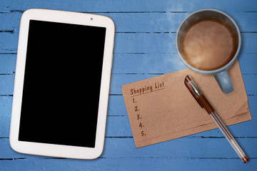 SHOPPING LIST ON NAPKIN PAPER WITH BLANK BLACK SCREEN TABLET HOT COFFEE AND A PEN ON BLUE OIL PAINTED WOODEN TABLE