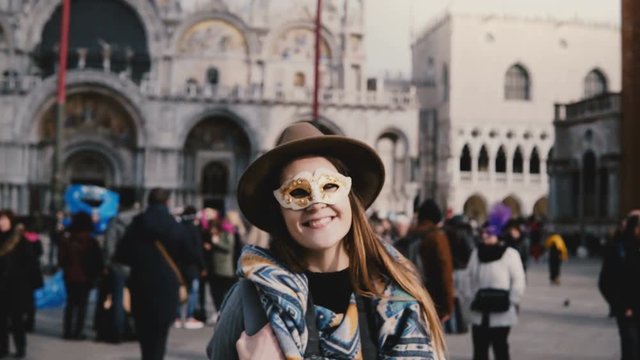 Portrait of happy woman with long hair wearing a white carnival face mask in Venice city square, Italy blowing kisses.