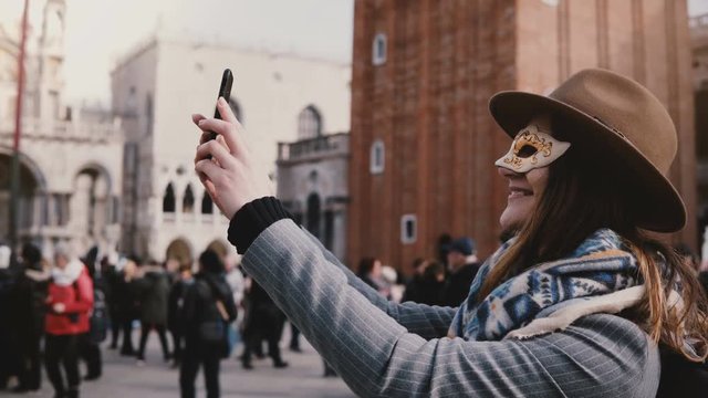 Happy woman wearing a stylish hat and white carnival face mask takes selfie photo smiling at Venice city square, Italy.