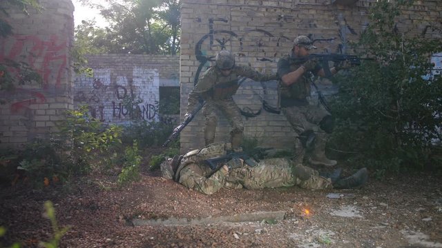 Army medic providing medical care to wounded in leg soldier while ranger securing area during combat. Military medic applying pressure bandage to casualty, binding gunshot wound, trying stop bleeding.