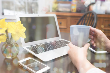 Naklejka na ściany i meble WOMAN WORKING FROM HOME (IN THE KITCHEN) HOLDING A CUP OF COFFEE SURROUNDING BY LAPTOP, SMART PHONE, TABLET, AND A VASE OF YELLOW DAFFODILS WITH NICE BOKEH OVER THE IMAGE. (WORK FROM HOME CONCEPT)