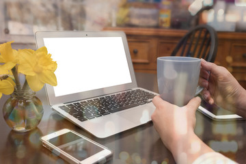 WOMAN WORKING FROM HOME (IN THE KITCHEN) HOLDING A CUP OF COFFEE SURROUNDING BY GADGETS AND A VASE OF YELLOW DAFFODILS. WHITE SCREEN FOR ADD TEXT, IMAGE, WEBSITE. (WORK FROM HOME CONCEPT)