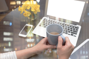 WOMAN WORKING FROM HOME (IN THE KITCHEN) HOLDING A CUP OF COFFEE WITH BLUR BACKGROUND OF GADGETS AND A VASE OF YELLOW DAFFODILS. WHITE SCREEN FOR ADD TEXT, IMAGE, WEBSITE. (WORK FROM HOME CONCEPT)