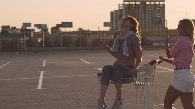 Fashion funny young hipster girls having fun at the shopping mall parking, roll each other in shopping cart on street. Slow motion