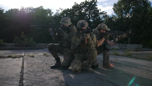 Elite squad of special forces rangers with combat rifles in camouflage keeping all-round defense, aiming weapons at enemy target outdoors. Army soldiers in defensive position during military operation