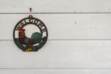 Old rusty metal rooster welcome sign hanging on white wood background.