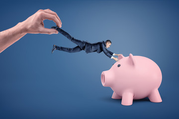 A tiny businessman tries to grab at a large piggy bank but he dragged away by a giant hand.