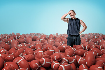 A thoughtful muscular man stands in a field of identical American football balls.