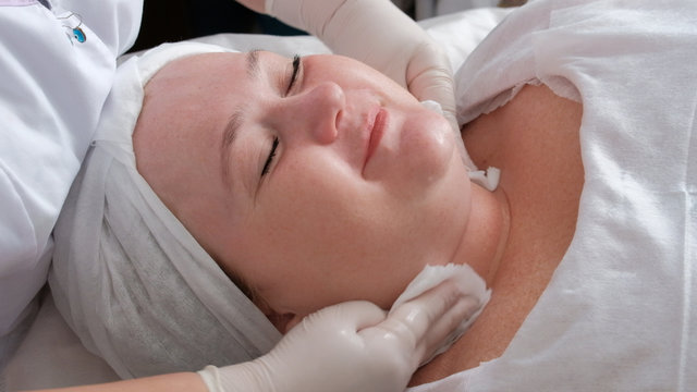 Cosmetological beauty procedure. Full woman smiling. The hands of a beautician in white gloves wipe the girl's face with wet wipes. Skin cleaning and aesthetic care in the cosmetology center.