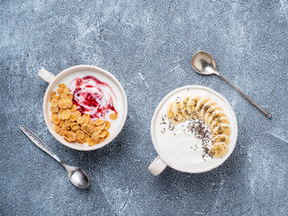 Two greek yogurt with jam, muesli, chia seeds and banana in white bowl on grey blue concrete stone table, top view