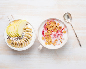 Two greek yogurt with jam, apple, muesli, chia seeds and banana in white bowl on white wooden table, top view