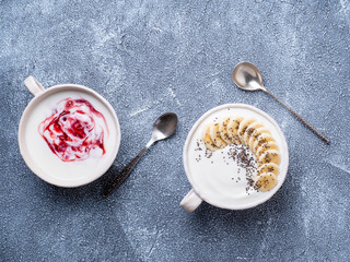 Two greek yogurt with jam, chia seeds and banana in white bowl on grey blue concrete stone table, top view
