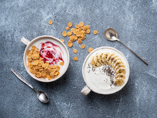 Two greek yogurt with jam, muesli, chia seeds and banana in white bowl on grey blue concrete stone table, top view