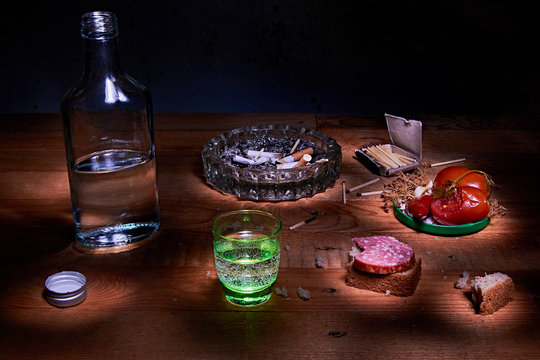 Still life with the concept of alcoholism. The composition consists of a bottle of vodka, a stack, an ashtray with cigarette butts and snacks.