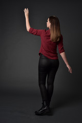 full length portrait of brunette girl wearing  red shirt and leather pants. standing pose, facing away from the camera, on grey studio background.