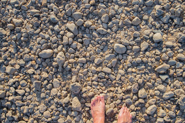 Naked bare feet stand on the ground with numerous pebbles.