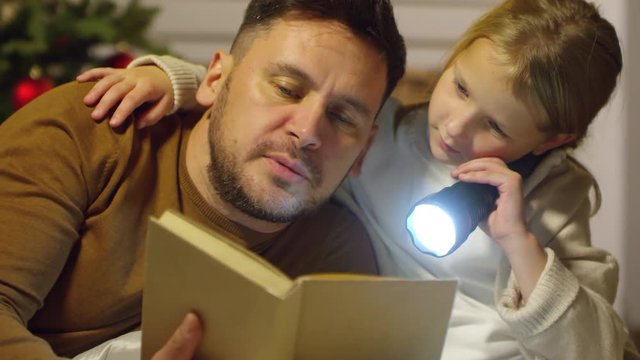Little girl holding flashlight, embracing father and listening to him while reading fairytale together in the evening on Christmas Eve