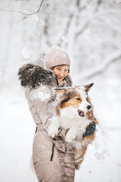 A Beautiful Young Woman In Warm Clothes Holding Her Beloved Dog In Her Arms And Laughing, A Lot Of Snow Will Fall From Above.
