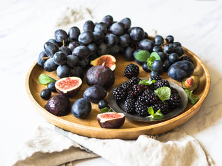 Purple fruits, berries and green mint leaves. Blackberries, grapes, plums and figs in a wooden tray on a white marble background. Tasty and ripe fruits and berries. Top view