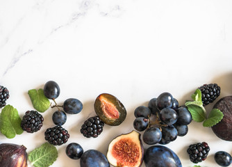 Purple fruits, berries and mint leaves border. Blackberries, grapes, plums and figs on white marble background. Tasty and ripe fruits and berries. Copy space. top view