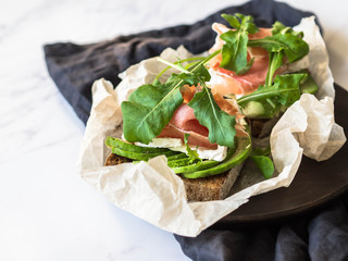 Toasts with camembert cheese, avocado slices, prosciutto, arugula and spices on wood plate on marble table. Copy space