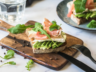 Toasts with camembert cheese, avocado slices, prosciutto, arugula and spices on wood board on marble table
