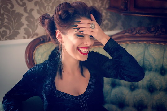 Sexy Caucasian model looking like a vampire laughing on a couch, having sculpting make-up and cocktail dress with big cleveage. Maleficent lookalike retro concept
