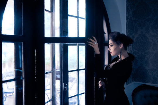 Sexy Caucasian model like a vampire looking through the window, having sculpting make-up and cocktail dress with big cleveage. Maleficent lookalike concept