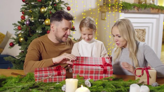 Happy mother and dad smiling and talking to little daughter while opening Christmas present together in the living room decorated with lights and fir tree