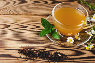 Full glass cup of green tea and mint and bunch of chamomile near scattered dry tea on old wooden table. Healthy concept