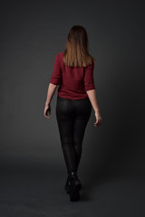 full length portrait of brunette girl wearing  red shirt and leather pants. standing pose, facing away from the camera, on grey studio background.