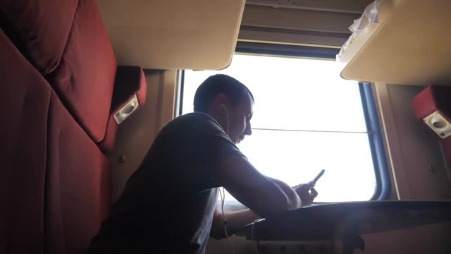 man silhouette travel is sitting on the train carriage holding sits by the window a smartphone Railway in headphones. slow motion video. man writes messages in the smartphone in the train social media