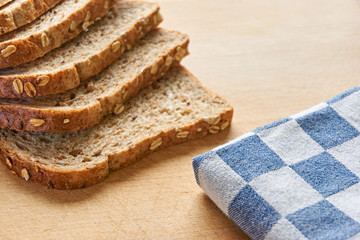 Close-up of slices of whole grain bread on a wooden cutting board with bran and a blue and white checkered cloth in the background
