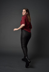 full length portrait of brunette girl wearing  red shirt and leather pants. standing pose, facing away from the camera, on grey studio background.