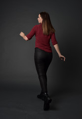 full length portrait of brunette girl wearing  red shirt and leather pants. standing pose, facing away from the camera, on grey studio background.