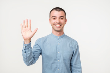 Attractive european student waives hand in hello gesture while smiling cheerfully over white background.