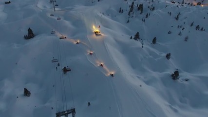 Aerial view of a snow groomer and snow mobiles on a ski slope in the French Alps. Sunset time low light. Snowy landscape.